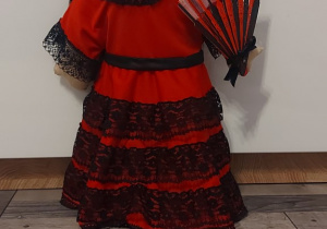 Laleczka Nadii Tułazy: Przedstawia lalkę hiszpankę. Długie czarne włosy wykonane z włóczki. Piękna długa, czerwona sukienka z czarną koronką. Naszyjnik wokół szyi również wykonany z koronki czarnej. Laleczka w lewej ręce trzyma czerwono-czarny wachlarz.